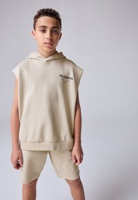 Felpa senza maniche beige con cappuccio e logo frontale, abbinata a pantaloncini coordinati. Il tessuto sembra morbido, con una texture liscia e un design minimalista.