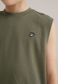 Olivgrön ärmlös t-shirt i mjuk bomull med en liten broderad fågel-logotyp på bröstet. Slät textur, rundad halsringning.