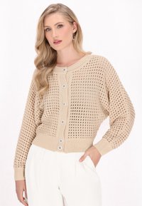 Cardigan lavorato a maglia beige con motivo a rete aperta, scollatura rotonda e cinque bottoni frontali. Polsini e orlo a coste, abbinato a pantaloni bianchi.