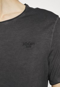 Primo piano di una persona che indossa una maglietta nera a girocollo con "JOOP! JEANS" ricamato sul lato sinistro del petto.