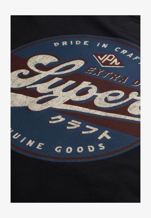 Camiseta navy con un logo redondo que presenta texto en blanco y rojo. El diseño incluye un patrón texturizado y caracteres japoneses sobre un fondo azul.