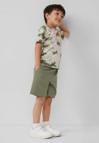 T-shirt a maniche corte verde tie-dye con un accento arancione, abbinato a pantaloni corti verde oliva e sneakers bianche con strap in velcro.
