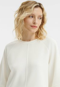 Maglione beige chiaro con collo rotondo, maniche raglan e una texture morbida. Presenta dettagli delle cuciture lungo il fronte per uno stile minimalista.