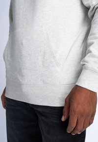Sudadera gris claro con bolsillo tipo canguro, puños y dobladillo acanalados, hecha de una tela suave y texturizada. Jeans negros visibles en la parte inferior.