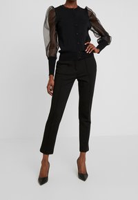 Cárdigan negro con mangas abullonadas transparentes y botones frontales, combinado con pantalones negros de corte ajustado y tacones puntiagudos negros. Texturas suaves en todo el conjunto.