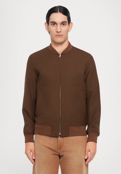 Veste bombardier marron avec fermeture éclair à l'avant, col et poignets côtelés. Comprend deux poches latérales et une texture lisse. Portée avec un pantalon beige.