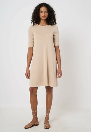 Kobieta o kręconych włosach, ubrana w beżową sukienkę do kolan z podwiniętymi rękawami oraz beżowe sandały na płaskiej podeszwie z paskami, stojąca na jednolitym tle.