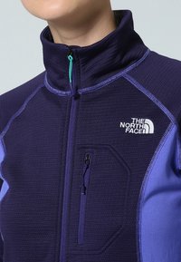 Polarowa kurtka w kolorze fioletowym z kontrastującymi jasnofioletowymi akcentami. Posiada wysoki kołnierz, zapinaną na zamek kieszeń na piersi oraz logo na klatce piersiowej.
