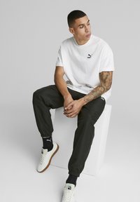 Hvid bomulds t-shirt med sort Puma logo, sorte joggerbukser og hvide sneakers med gummisål. Sidder i en pose på en hvid kube.