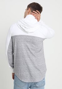 Grå och vit huoodie med raglanärmar, slät textur och förlängd bakre fåll. Har en dragsko för huvan.