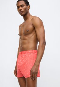 Κόκκινα μαγιό σορτς με διακριτικό λευκό μοτίβο λογοτύπου, ελαστική μέση και χαλαρή εφαρμογή, κατασκευασμένα από ελαφρύ, γρήγορα στεγνάζον ύφασμα.