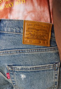 Gros plan sur un jean Levi's en denim bleu montrant un patch en cuir avec le logo et une étiquette rouge Levi's sur la poche arrière, porté avec une chemise rouge.