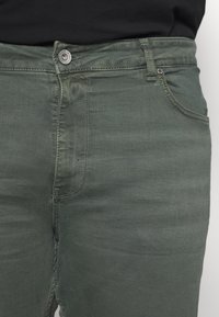 Green denim jeans with a slim fit, featuring a button closure, front pockets, and contrasting stitching on the seams.