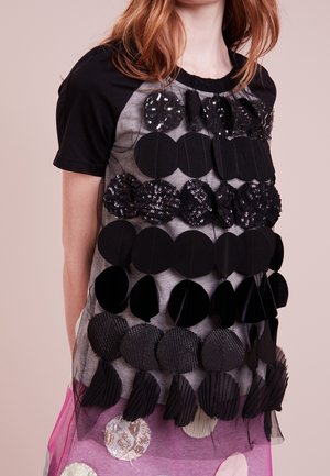 Kobieta ubrana w czarną bluzkę z krótkim rękawem ozdobioną warstwami okrągłych czarnych i cekinowych aplikacji na przezroczystej fioletowej spódnicy z dużymi łatami.