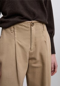 Pantalones beige con tela texturizada, frente con pliegues y cierre con un solo botón. Combinados con un suéter marrón, mostrando la zona de la cintura.