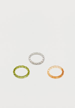 Tri prstena od perli u srebrnoj, prozirno zelenoj i prozirno jantarnoj boji, postavljena na bijeloj pozadini.