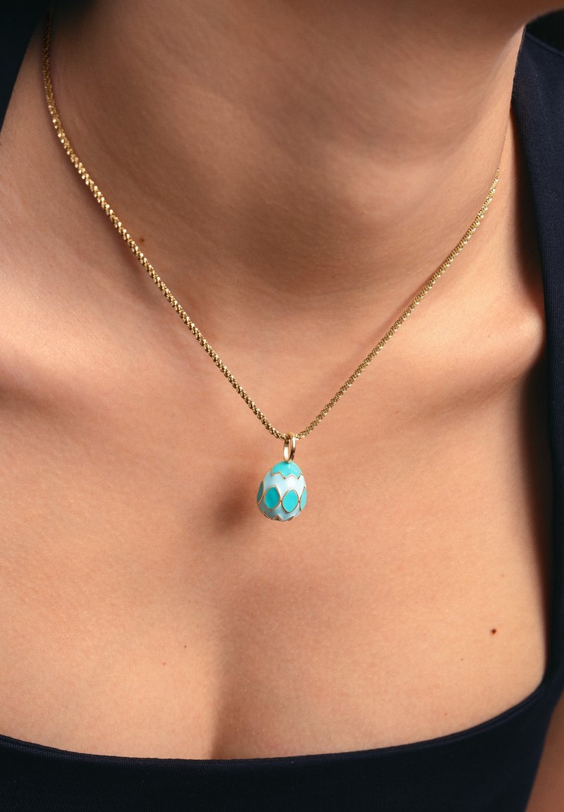 Collana in oro con ciondolo a forma di uovo turchese con dettagli bianchi e dorati, appoggiata su un collo dalla tonalità della pelle.