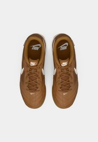 Smeđe Nike tenisice s teksturiranim gornjim dijelom, bijelim Swoosh logotipom i smešnim vezicama. Potplat ima gumenu obradu i složene detalje šivanja.