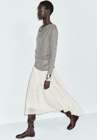 Maglia grigia a maniche lunghe con scollo drappeggiato, abbinata a una gonna bianca fluida e stivali di stoffa marroni al ginocchio. Bracciale a maniche in argento come accessorio.