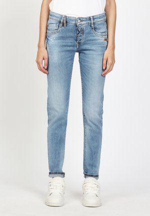 Woman wearing light blue slim-fit jeans and white sneakers, standing against a plain white background.