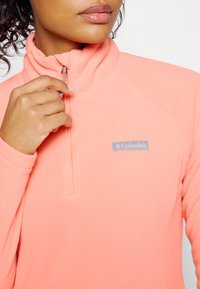 Hand van vrouw die een koraalroze Columbia fleece sweatshirt met hoge kraag aan het ritsen is, waarbij het onderste deel van het gezicht en de textuur van de stof zichtbaar zijn.