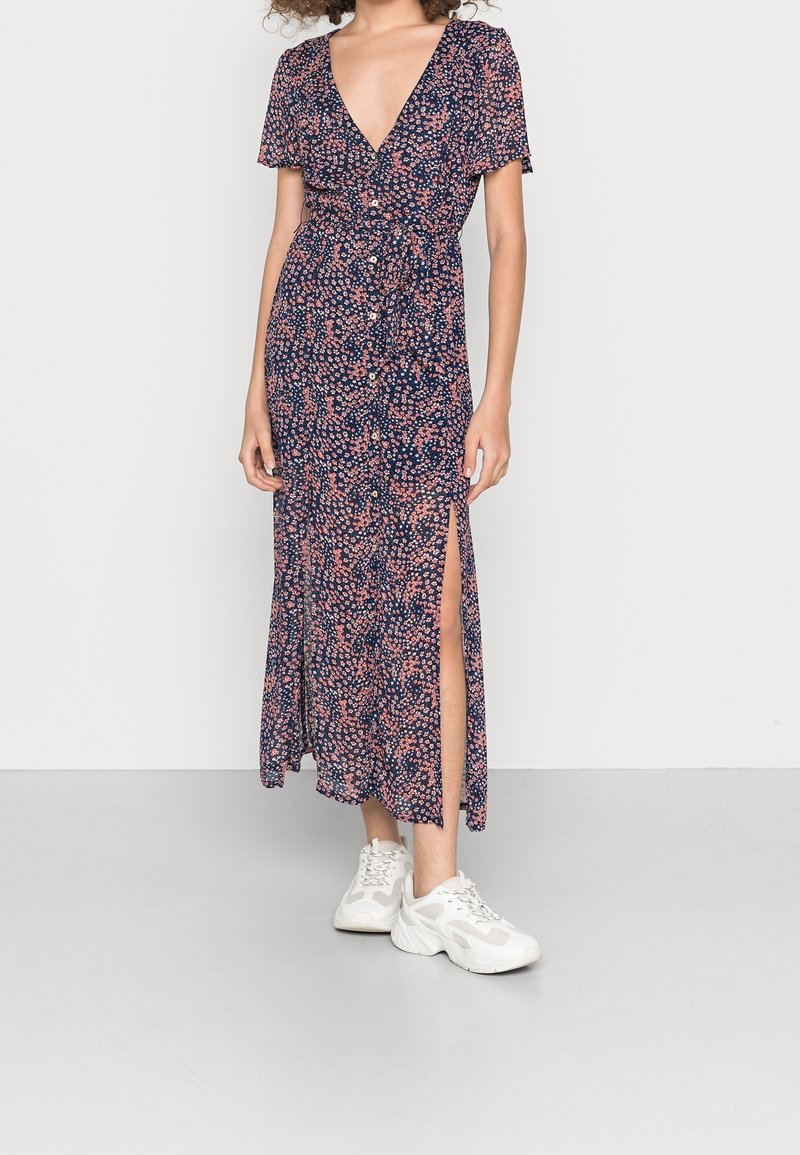 Abito midi floreale blu scuro con maniche corte, scollo a V e spacchi laterali. Indossato con sneakers bianche, presenta una vita regolabile con lacci e dettagli con bottoni.