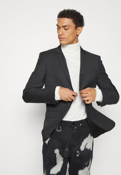 Dunkelgrauer Blazer mit tailliertem Design, Ein-Knopf-Verschluss, mit Revers, getragen über einem weißen Rollkragenpullover und schwarzen gemusterten Hosen.
