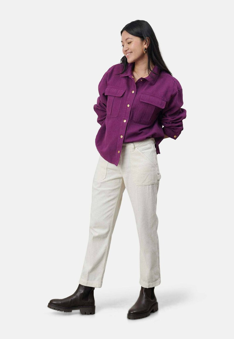 Fioletowa koszula z guzikami, z kieszeniami na piersi, połączona z białymi dopasowanymi spodniami z prążkowanego materiału i czarnymi botkami za kostkę. Gładki materiał i dobrze dopasowany krój.