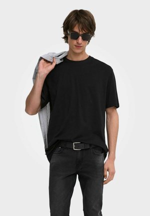 Giovane uomo con t-shirt nero e occhiali da sole tiene una giacca a righe sulla spalla davanti a uno sfondo semplice.