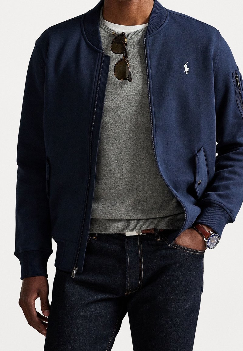 Homme portant une veste bomber bleu marine par-dessus un pull gris, jean foncé, lunettes de soleil accrochées au col du pull et montre-bracelet au poignet gauche.