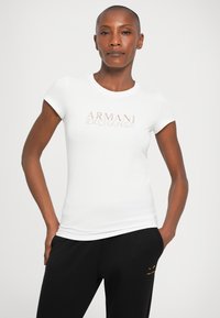 Biely tričko s krátkym rukávom a okrúhlym výstrihom, s logom "ARMANI EXCHANGE" v metalickej ružovej zlate. Bavlnený materiál, priliehavý strih.