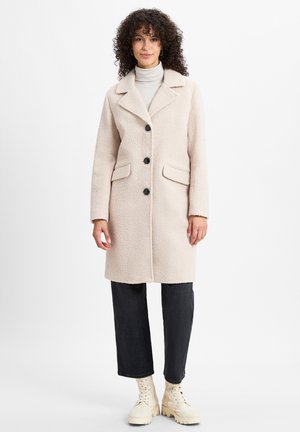 Beige textured coat with a notched collar, three black buttons, and side pockets, paired with dark jeans and cream boots.