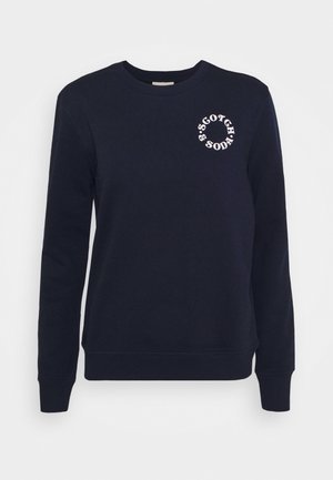 Granatowa bluza z długim rękawem i okrągłym dekoltem, z białym okrągłym napisem „Scotch & Soda” na górnej lewej części klatki piersiowej.