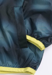 Veste légère avec un motif à pois bleu marine et vert, dotée d'une bordure jaune et d'une petite poche. Matière lisse et résistant à l'eau.