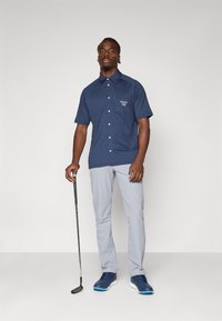 Marineblå kortermet skjorte med knapper og brystlomme, kombinert med lysegrå bukser og blå joggesko. Golfkølle holdt i høyre hånd.
