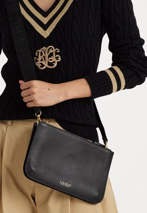 Personne portant un pull noir à encolure en V avec des rayures beige et un logo brodé, tenant un sac bandoulière en cuir noir Lauren Ralph Lauren.