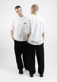 White oversized t-shirt features a small logo on the chest. Black loose pants with textured patches depict various designs. Black footwear.