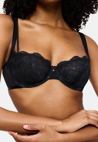 Γυναίκα φοράει μαύρο δαντελένιο σουτιέν με διπλές τιράντες, με τα χέρια σταυρωμένα κάτω από το στήθος.