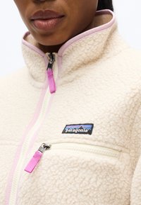 Κοντινό πλάνο ενός ατόμου που φοράει ένα κρεμ φλις μπουφάν Patagonia με ροζ φερμουάρ στο φερμουάρ και στο στήθος.