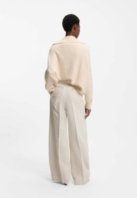 Person med kort lockigt hår som bär en lös krämfärgad tröja och vida beige byxor, står med händerna i fickorna och vänder ryggen mot oss.