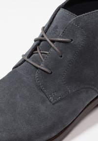 Bottine en daim gris avec une texture lisse, munie de lacets fins et de détails cousus sur les côtés. La semelle semble durable et offrant un bon soutien.