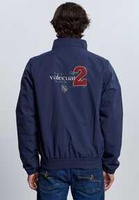 Chaqueta bomber azul marino con puños y dobladillo elásticos. Cuenta con un logo bordado, "Est. 1971," "valecuato2," y un diseño de escudo en la parte trasera.