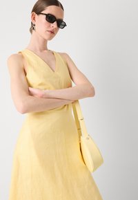 Rochie galbenă din in, cu decolteu în V și design fără mâneci, asortată cu o poșetă galbenă mică și ochelari de soare negri. Textură netedă, detalii minime.