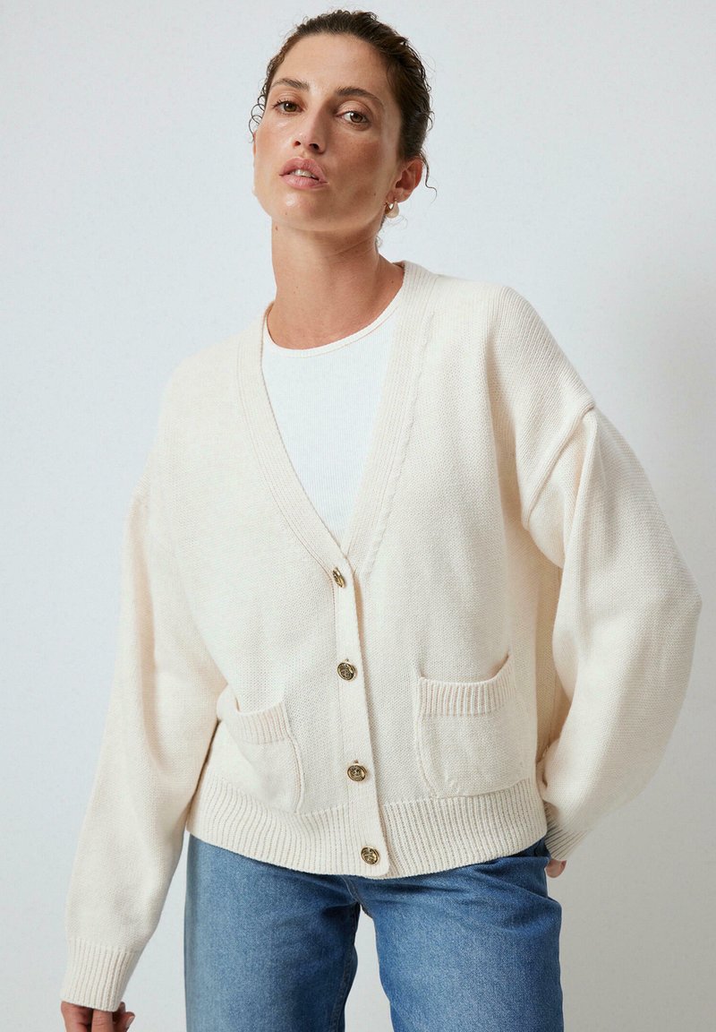 Crème kleurige cardigan met een V-hals, vier knopen aan de voorkant, textuur stof en twee zakken aan de voorkant. Gepaard met lichtblauwe jeans.