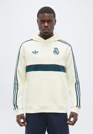 Hombre con sudadera con capucha crema con una franja horizontal verde oscuro, logo de Adidas y escudo del Real Madrid, posando frente a un fondo blanco liso.