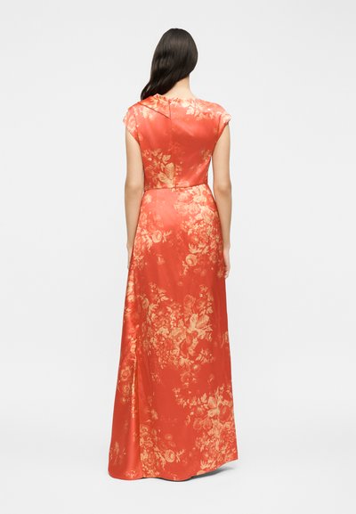 Γυναίκα με μακριά σκούρα μαλλιά, ντυμένη με ένα κοραλί φόρεμα μέχρι το πάτωμα με κοντά μανίκια και χρυσά λουλουδένια σχέδια, στέκεται γυρισμένη με την πλάτη σε ένα απλό φόντο.