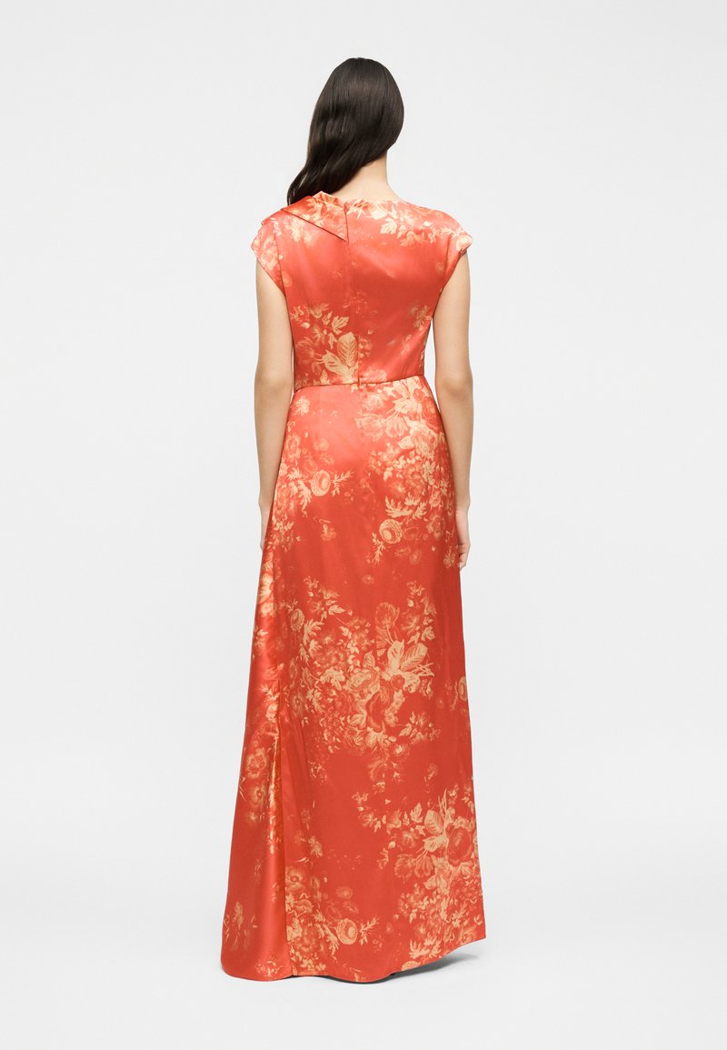 Woman with long dark hair wearing a floor-length, short-sleeve coral dress with gold floral patterns, standing facing away on a plain background.