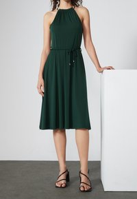 Abito verde senza maniche con scollo a halter arricciato, vita cinte con cordoncino e gonna fluttuante a campana. Indossato con tacchi neri a fasce.