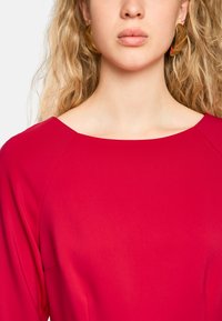 Rotes Kleid mit weitem Ausschnitt und nahtlosem Schulterdesign. Das Material ist glatt, und der Stoff hat einen leichten Glanz. Goldene Creolen sind sichtbar.
