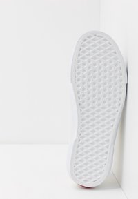 Vit sneaker med en texturerad gummisula som har ett diamantmönster. Ovandelen har en slät yta och minimalistiska designinslag.